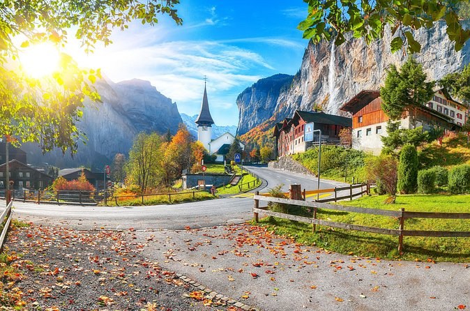 An autumn road with fallen leaves leading to a church spire under towering cliffs