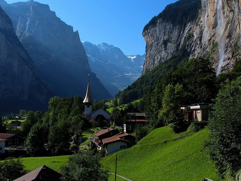 A white church and chalets under a tall waterfall pouring off a sheer cliff