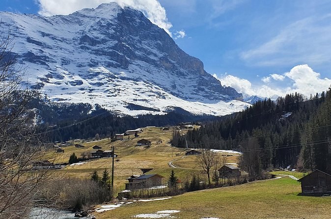 A huge snow-streaked mountain face above brown spring meadows and scattered huts