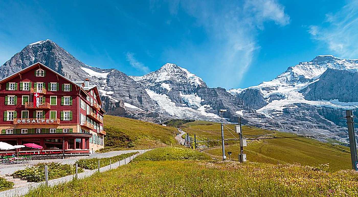 Three snow peaks in a row behind a red mountain hotel above a flowering meadow